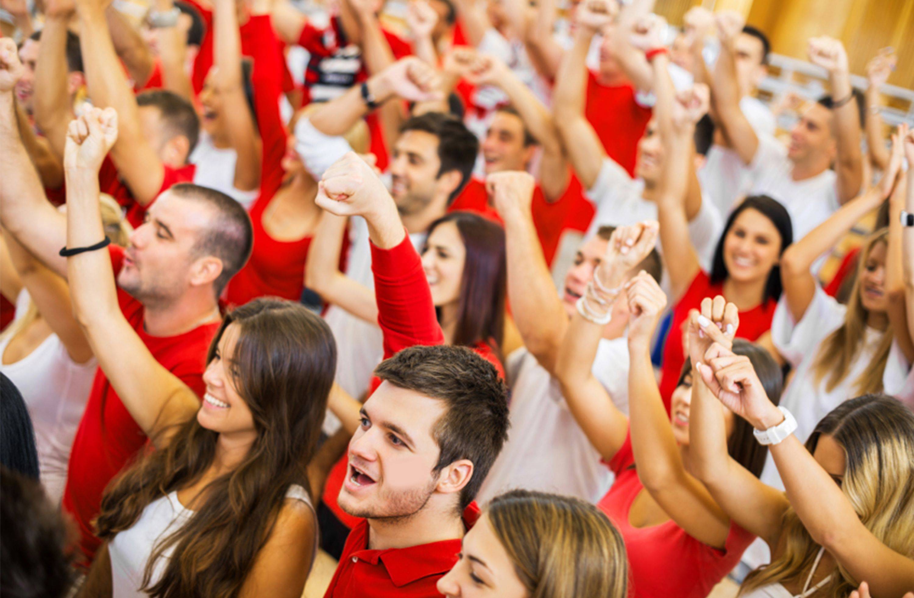 A zoomed-in photo of young adults wearing red and white shirts. They are all raising their arms in the air and making fists with their hands.