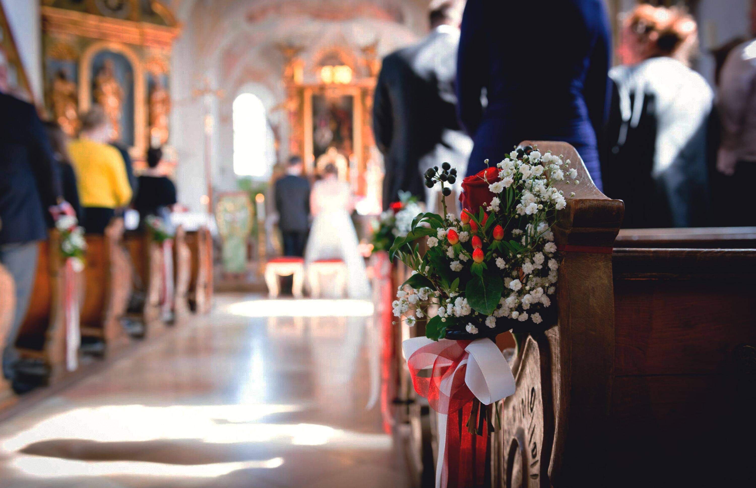 A depiction of a wedding with a bride and groom at the front. In the middle of the aisle, on the end of a pew, there's a flower arrangement in focus.