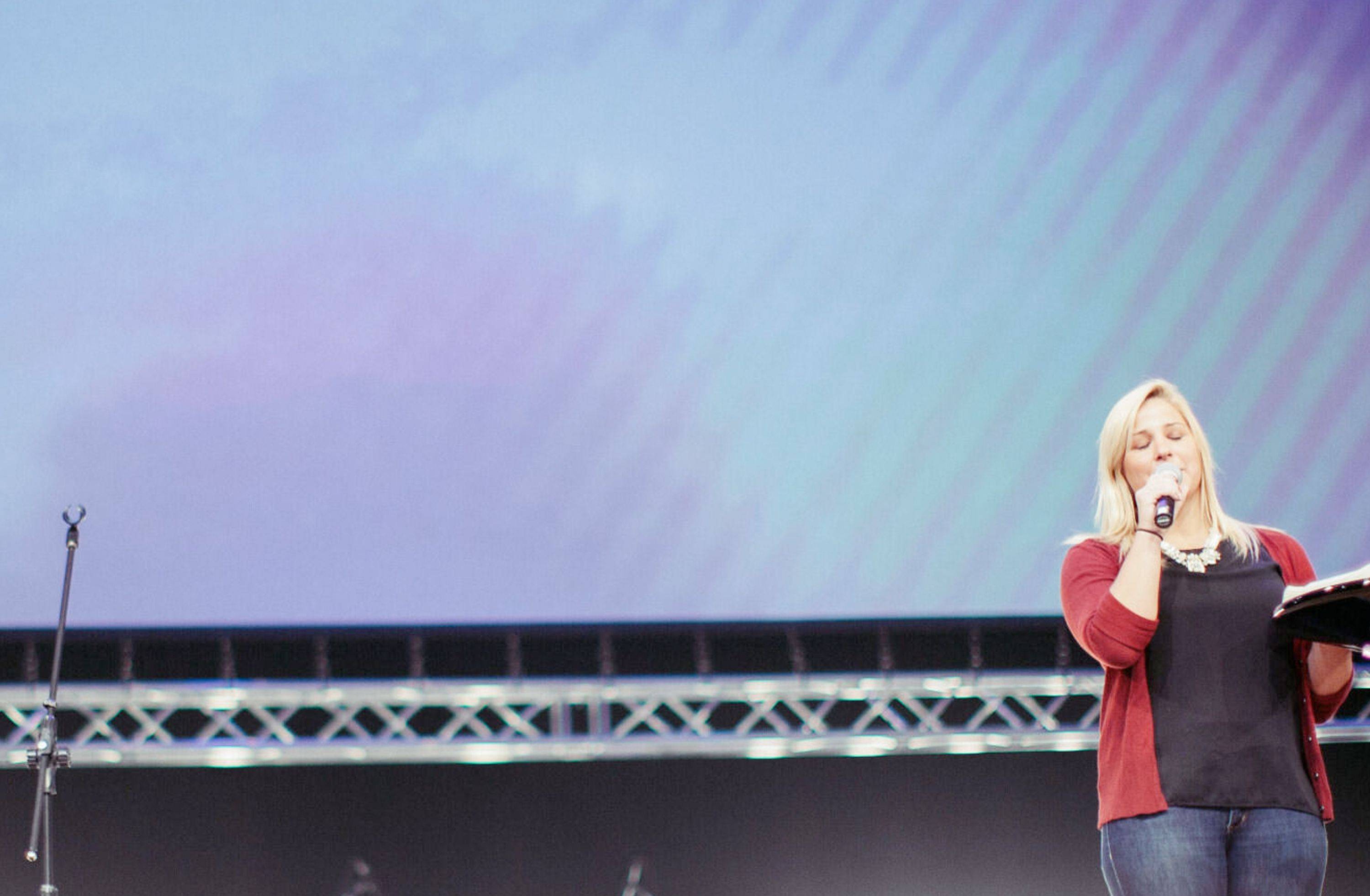 A zoomed-in perspective from the front row of a church setting. There's a person with their eyes closed holding a microphone to their mouth while standing at the pulpit. A large projection is on the wall behind them.