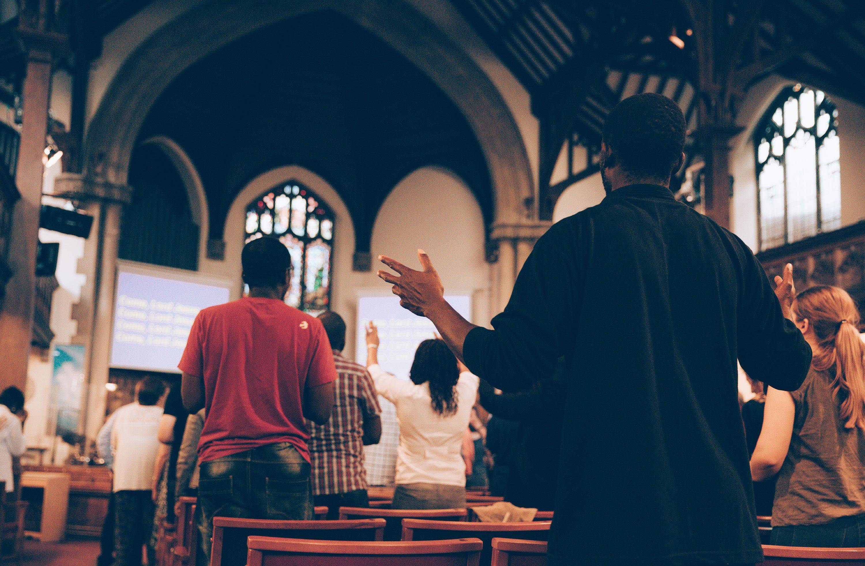 From the perspective of someone sitting down during worship, you see the backs of a dozen people as they engage in worship. In the distance you can see a stage with words being displayed on two screens on the left and right of the stage.